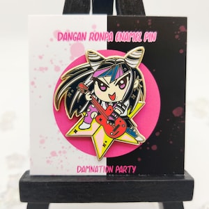Op de afbeelding: Een Dangan Ronpa emaille pin met een stripfiguur die een rode gitaar bespeelt. De pin is geplaatst tegen een roze cirkel en een stervormige achtergrond. De tekst "Dangan Ronpa Enamel Pin" en "Damnation Party" zijn zichtbaar.
