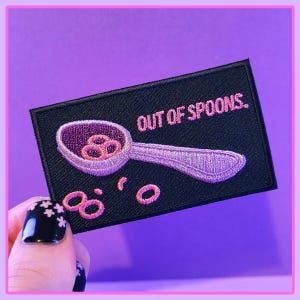 May include: A black embroidered patch with pink stitching. The patch features a spoon with pink cereal rings inside and the text "OUT OF SPOONS."