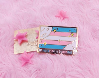 May include: Gold-tone enamel pin featuring a transgender pride flag with a crescent moon, stars, and clouds. The text "my pride is beautiful" is written below the flag.