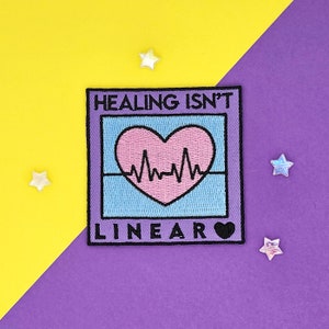 May include: A purple embroidered patch with a pink heart with a heartbeat line inside. The patch has a blue background and a black border. The text "HEALING ISN'T LINEAR" is written in black letters on the patch.