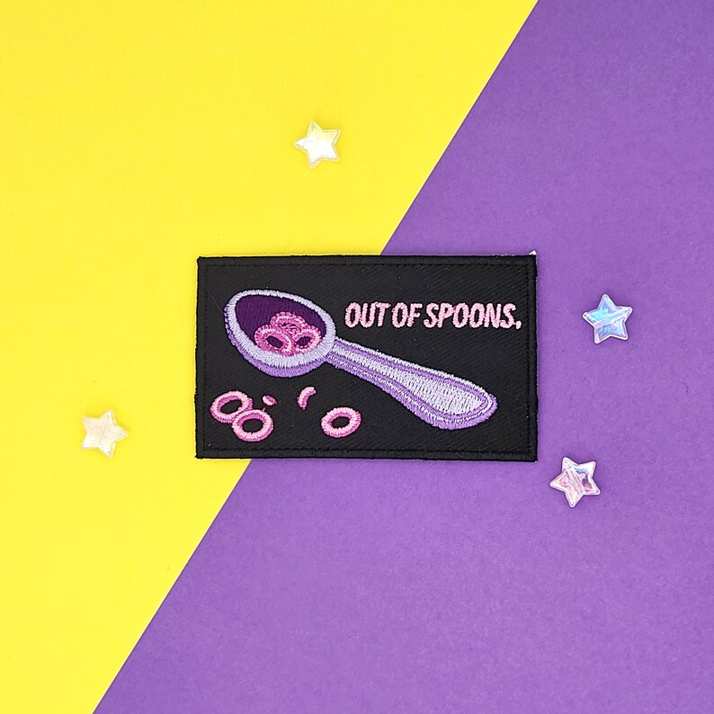 Puede incluir: Un parche bordado negro con una cuchara blanca llena de anillos de cereal rosados y el texto "OUT OF SPOONS."