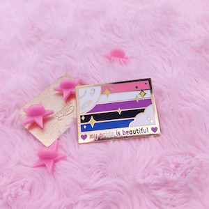 Pode incluir: Um broche de esmalte dourado com um design de bandeira multicolorido e o texto "my pride is beautiful". A bandeira tem listras rosa, brancas, roxas, pretas, azuis e brancas com estrelas douradas e nuvens. O broche está sobre uma superfície rosa e felpuda.