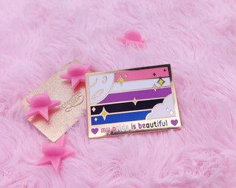 May include: A gold-tone enamel pin with a multi-colored flag design and the text "my pride is beautiful". The flag has pink, white, purple, black, blue, and white stripes with gold stars and clouds. The pin is on a pink fuzzy surface.
