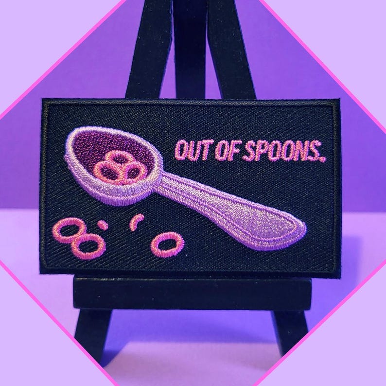 Puede incluir: Un parche bordado negro con puntadas rosas. El parche presenta una cuchara con anillos de cereal rosas que se derraman. El texto "OUT OF SPOONS." est&aacute; bordado en rosa en el parche.