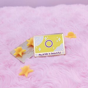 Pode incluir: Um broche de esmalte dourado com fundo amarelo e um círculo roxo com contorno branco. O broche apresenta uma nuvem branca, estrelas e o texto "my pride is beautiful".