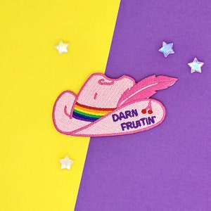 May include: Pink cowboy hat with a rainbow band and a feather. The hat has the text "DARN FRUITIN" on it.