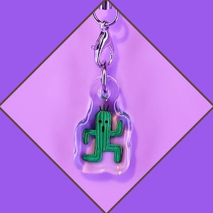 Könnte beinhalten: Ein durchsichtiger Acryl-Schlüsselanhänger mit einer grünen Kaktusfigur im Inneren. Der Schlüsselanhänger hat einen silbernen Metallverschluss.