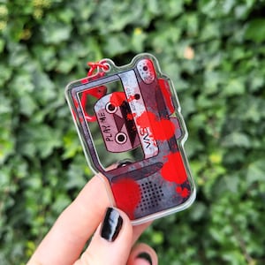 May include: A clear acrylic keychain in the shape of a cassette tape, with a red and grey design and blood splatter details. The text "PLAY ME" is visible, along with the letters "VAS". A red loop is attached for hanging.