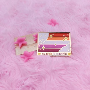 May include: A gold-toned enamel pin with a multi-coloured striped design and stars. The pin features a pink, red, orange, yellow, and purple striped design with a white cloud and a crescent moon. The text "my pride is beautiful" is written below the design.