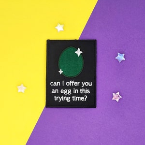 Op de afbeelding: Zwarte geborduurde patch met een groen ei en witte sterren. De tekst luidt "Kan ik je in deze moeilijke tijd een ei aanbieden?"