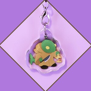 Könnte beinhalten: Ein durchsichtiger Acryl-Schlüsselanhänger mit einer Comicfigur, die ein Messer und eine Laterne hält. Die Figur ist grün, braun und gelb. Der Schlüsselanhänger hat einen silbernen Metallverschluss.