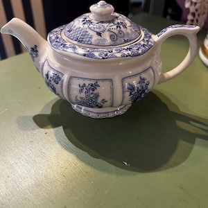 May include: A white ceramic teapot featuring a blue floral pattern with intricate scrollwork and geometric borders. The teapot has a curved handle, a tapered spout, and a matching lid with a small knob handle, all decorated in blue.