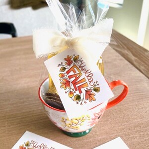 May include: A white ceramic mug with a floral design and a red handle. The mug is filled with a clear liquid and a chocolate cookie. A white gift tag with the text "Hello Fall" and a fall-themed design is attached to the mug.