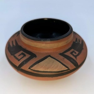 May include: A handcrafted, terracotta-colored pottery bowl with a black interior. The exterior features black geometric designs and a tan triangle. The bowl has a wide, flat base and a slightly flared rim.