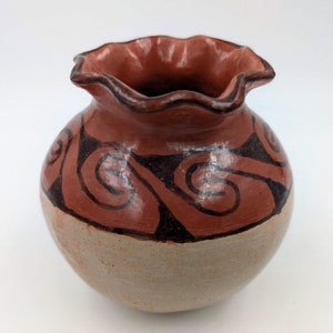 May include: A small, round, terracotta pottery vase with a scalloped rim. The vase is decorated with a brown and black geometric pattern.