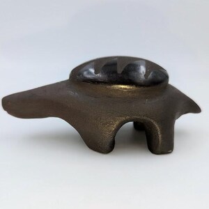 May include: A bronze-colored, abstract sculpture with a rounded top and a body supported by three legs. The sculpture has a smooth, metallic finish and is set against a white background. The design is minimalist and modern.