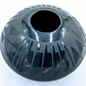May include: A black ceramic vase with a round shape and a wide opening. The vase features a geometric design in a lighter shade of black, creating a contrast. The design appears to be etched or painted onto the surface. The vase is likely used for decorative purposes.