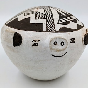 May include: A white ceramic pig-shaped bowl with a black and white geometric design on the top. The bowl has a smiling face with small eyes, a snout, and ears. The bowl is a decorative item.