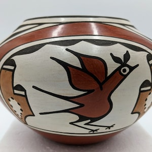 May include: A hand-painted pottery bowl with a red, white, and black design. The bowl features a stylized bird with a red body and black wings. The bird is flying in front of two brown and white geometric shapes.