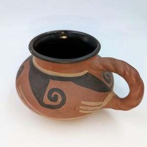 May include: A white ceramic mug with a black handle and the text "But First, Coffee" in a black, bold font. The mug is on a white surface.