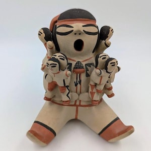 May include: A hand-painted ceramic figurine of a Native American woman with multiple children on her back. The figurine is painted in brown, black, red, and white.