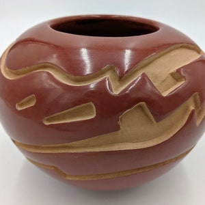 May include: A red clay pot with a carved design. The pot is decorated with a pattern of wavy lines and geometric shapes.