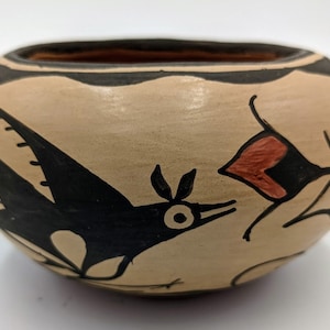 May include: A small, hand-painted pottery bowl with a black bird design and a red flower. The bowl is decorated with black and white lines and has a beige background.
