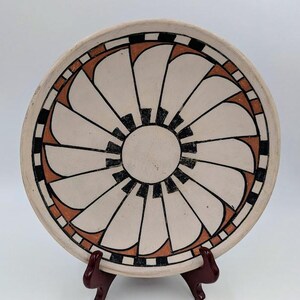 May include: A round, cream-colored ceramic plate with a geometric design. The design features a central black circle surrounded by white, black, and orange segments. The plate is displayed on a dark brown stand.