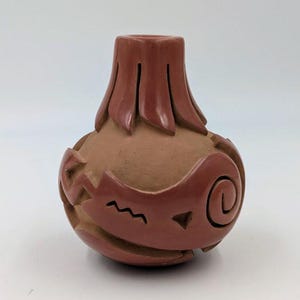 May include: A small, round terracotta vase featuring a matte finish and a polished, dark reddish-brown neck and base. The body is decorated with carved geometric patterns, including a spiral, a zigzag line, and a triangle, set against a tan surface.