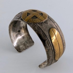 May include: A wide cuff bracelet made of tarnished silver-tone metal featuring an embossed texture. The bracelet is accented with a circular gold-tone medallion at the top and a smooth, elongated gold-tone feather-shaped inlay on the side.