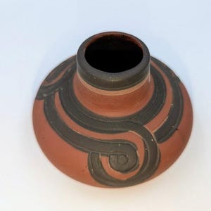 May include: A terracotta-colored ceramic vase with a dark brown interior and a decorative black swirl design. The vase has a wide, flat base and a narrow neck. The design is symmetrical and wraps around the vase.