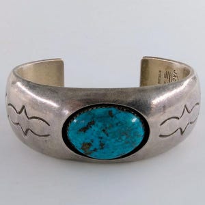 May include: A sterling silver cuff bracelet featuring a central oval turquoise stone with dark matrix. The metal band has a smooth, polished finish with etched geometric patterns on both sides. The interior is stamped with JAC and STERLING.
