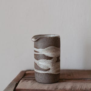 May include: A handmade ceramic pitcher with a spout, featuring a textured brown and cream wave pattern. The stoneware pitcher sits on a rustic wooden surface, showcasing its artisanal design and neutral color palette.