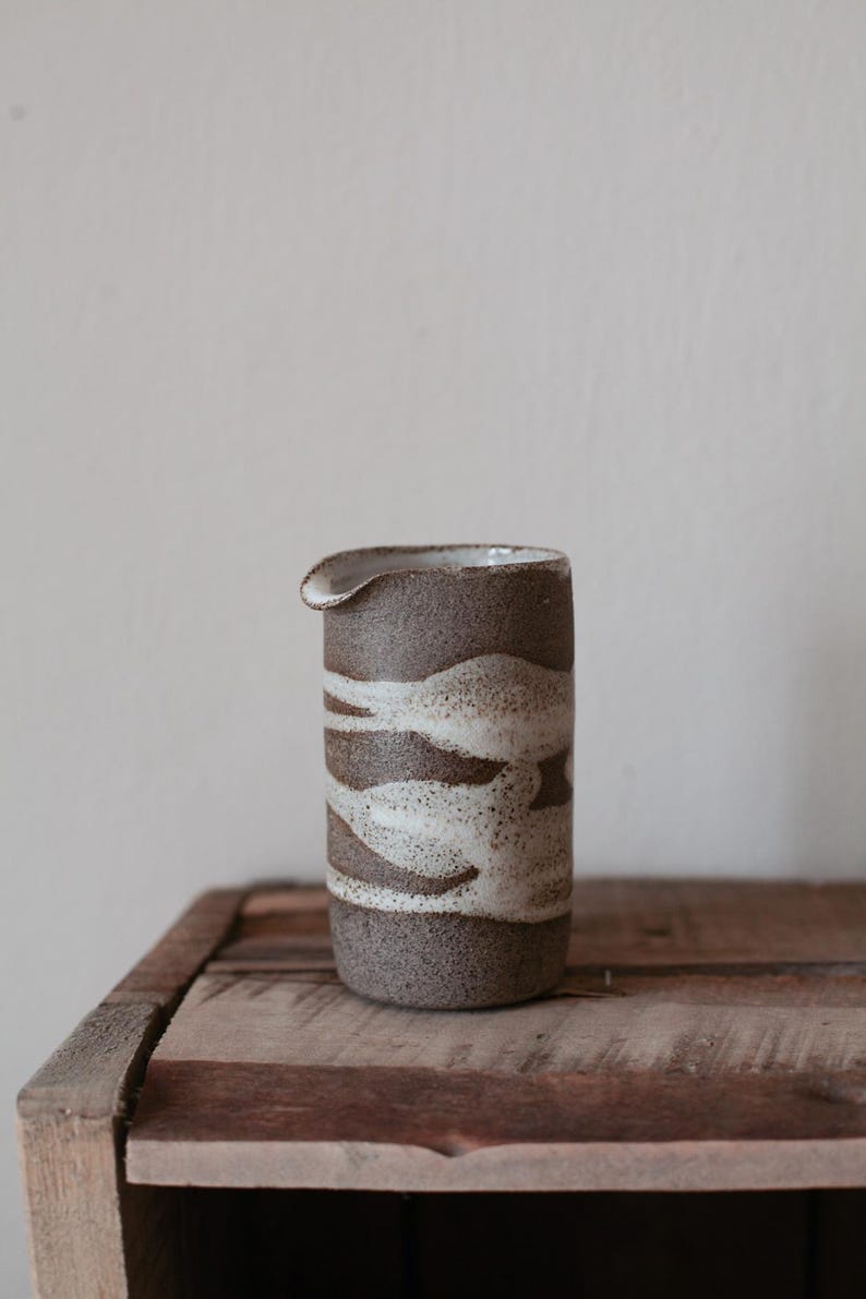May include: A handmade ceramic pitcher with a spout, featuring a textured brown and cream wave pattern. The stoneware pitcher sits on a rustic wooden surface, showcasing its artisanal design and neutral color palette.