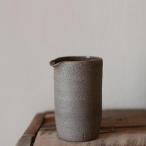 May include: A cylindrical, gray ceramic pitcher with a spout. The pitcher has a textured surface and a dark rim. It sits on a wooden surface. The pitcher is approximately 6 inches tall.