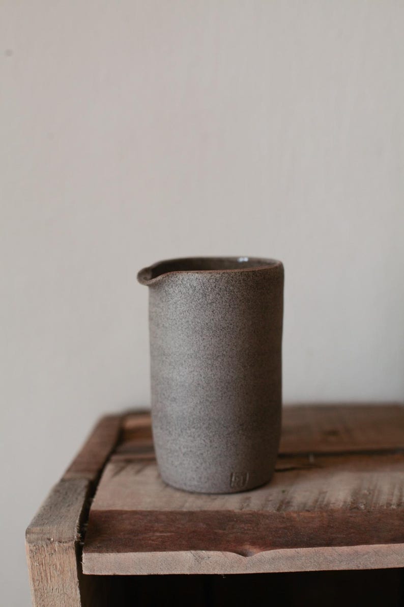 May include: A cylindrical, gray ceramic pitcher with a spout. The pitcher has a textured surface and a dark rim. It sits on a wooden surface. The pitcher is approximately 6 inches tall.