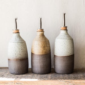 Könnte beinhalten: Drei Keramik-Ölflaschen mit Edelstahl-Ausgießern und Korken. Die Flaschen sind zweifarbig, mit einem dunkelbraunen Sockel und einem helleren, gesprenkelten oberen Bereich. Sie stehen auf einer rustikalen Holzoberfläche.