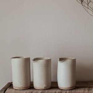 May include: Three off-white ceramic pitchers with a speckled texture and a small spout, arranged on a rustic wooden shelf. The pitchers have a minimalist design and a natural, earthy aesthetic. Dried flowers are in the background.