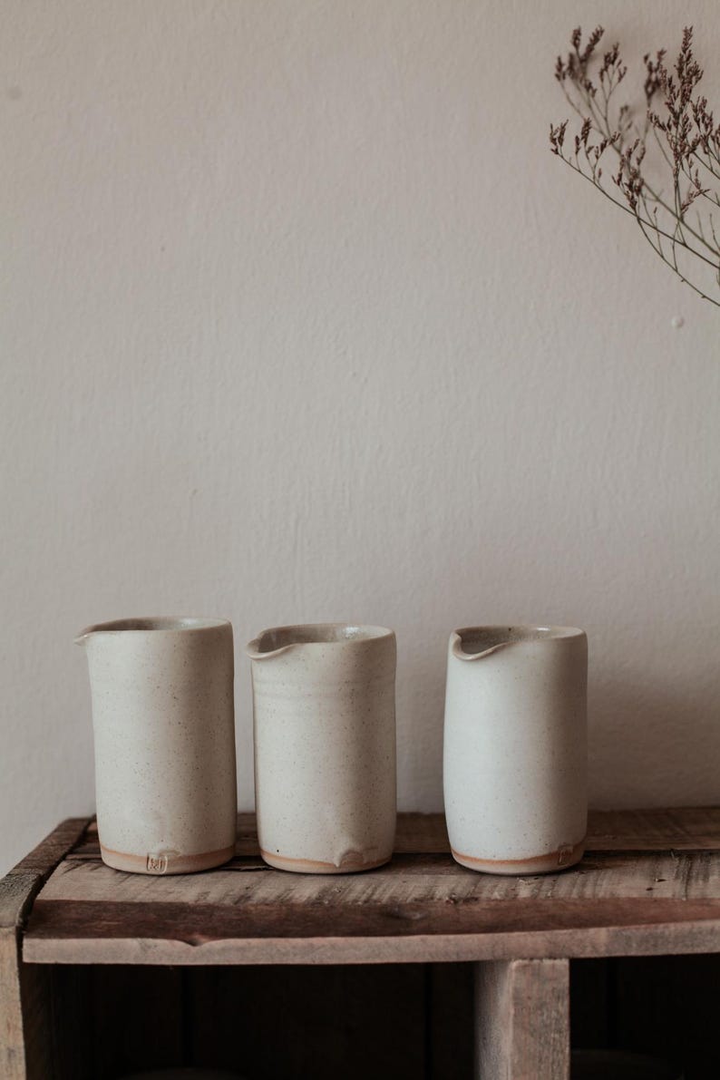 May include: Three off-white ceramic pitchers with a speckled texture and a small spout, arranged on a rustic wooden shelf. The pitchers have a minimalist design and a natural, earthy aesthetic. Dried flowers are in the background.