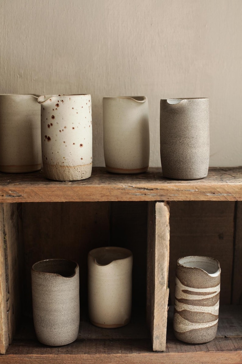May include: A collection of handmade ceramic pitchers in various neutral tones, including beige, speckled white, and gray. The pitchers are displayed on a rustic wooden shelf, showcasing their unique shapes and textures. The pitchers have a small spout.