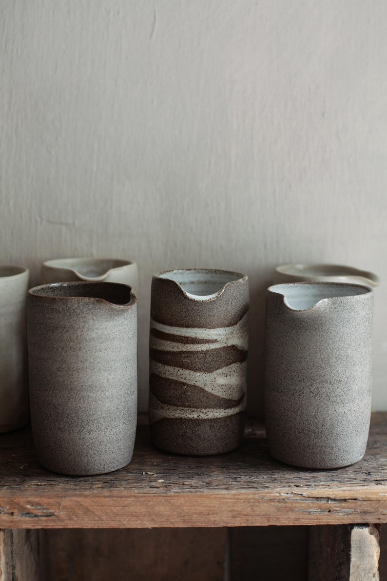 May include: A collection of handmade ceramic pitchers in various shades of gray and brown. The pitchers have a unique, organic shape with a spout-like opening. The textured surface adds to their rustic charm, perfect for a minimalist home decor.