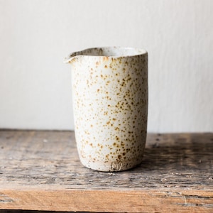 May include: A speckled ceramic creamer with a spout. The off-white vessel has brown spots and sits on a wooden surface. The creamer is cylindrical with a slightly irregular rim.