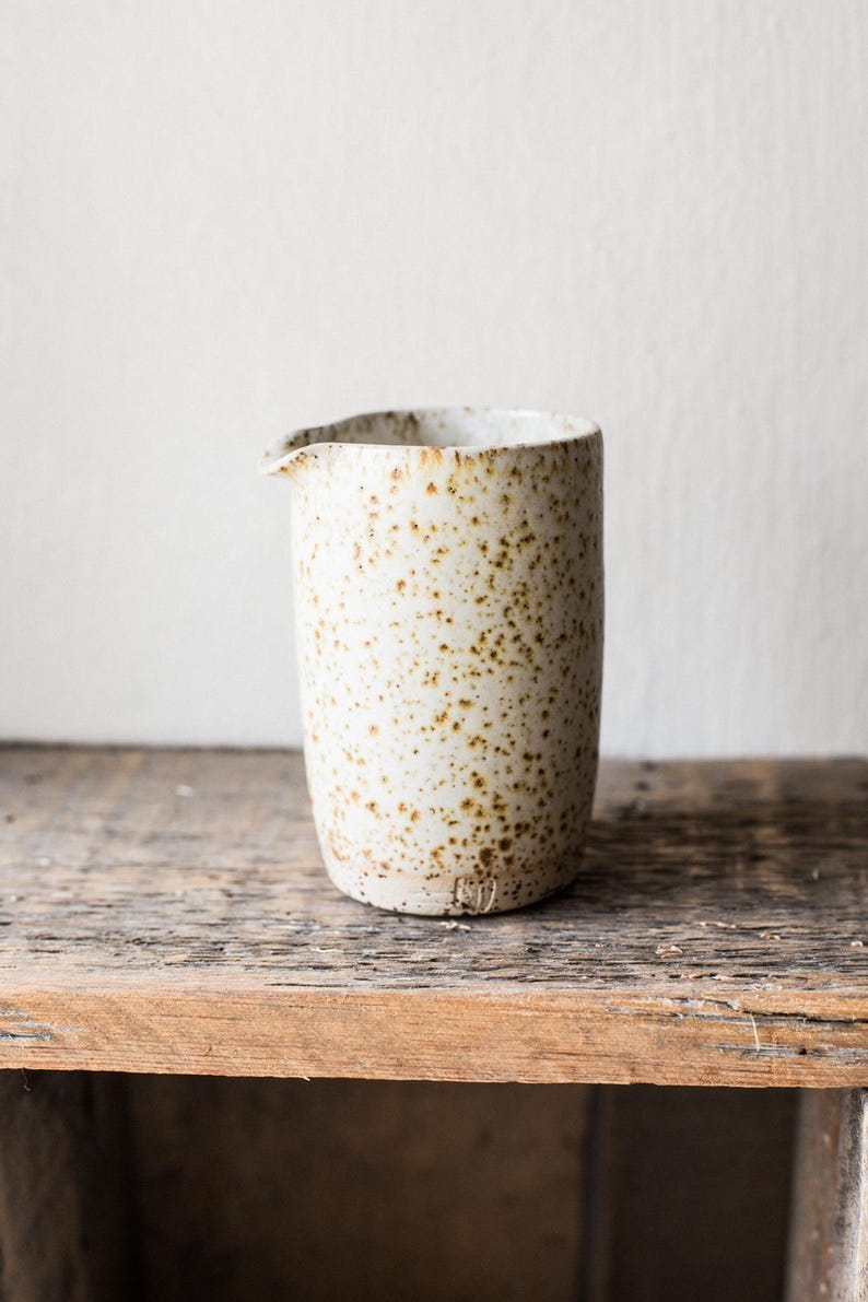 May include: A speckled ceramic creamer with a spout. The off-white vessel has brown spots and sits on a wooden surface. The creamer is cylindrical with a slightly irregular rim.
