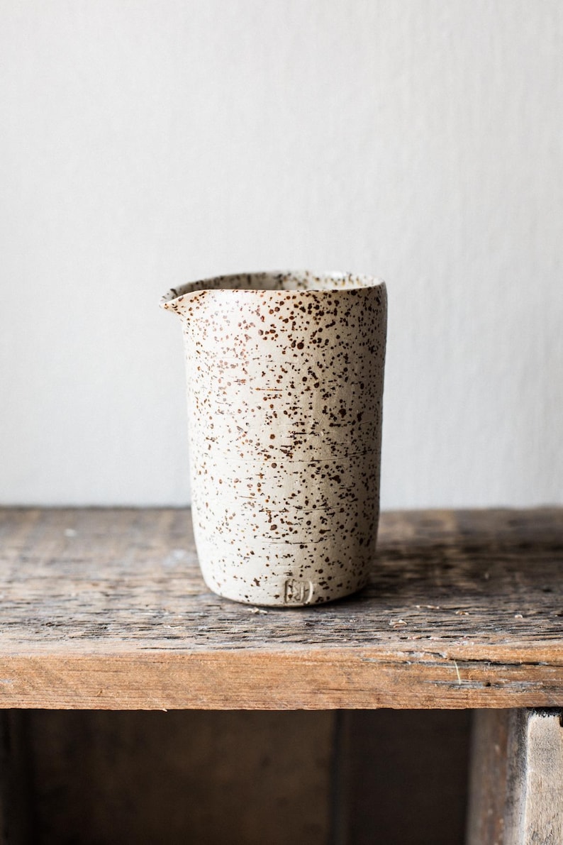 May include: A speckled ceramic pitcher with a spout, in a neutral color palette. The off-white body is covered in brown speckles. The pitcher sits on a wooden surface, suggesting a rustic or farmhouse aesthetic. The design is simple and functional.