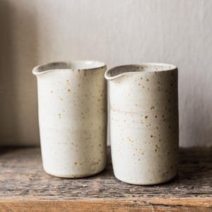 May include: Two off-white ceramic pitchers with a speckled finish. The pitchers have a small spout and are cylindrical in shape. They are displayed on a rustic wooden surface, with a neutral background.