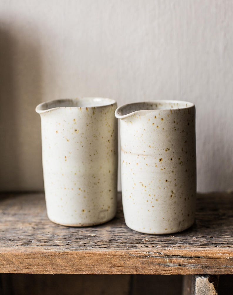 May include: Two off-white ceramic pitchers with a speckled finish. The pitchers have a small spout and are cylindrical in shape. They are displayed on a rustic wooden surface, with a neutral background.