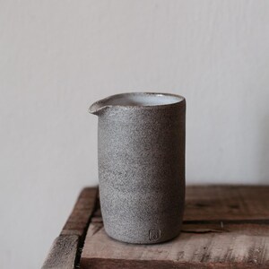 May include: A speckled gray ceramic pitcher with a spout, set on a rustic wooden surface. The cylindrical pitcher has a small, rounded spout and a textured surface. The interior is white. A small, square emblem is visible near the base.