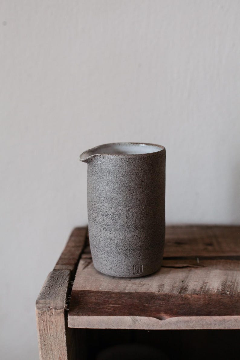 May include: A speckled gray ceramic pitcher with a spout, set on a rustic wooden surface. The cylindrical pitcher has a small, rounded spout and a textured surface. The interior is white. A small, square emblem is visible near the base.