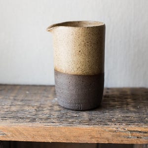 May include: A handmade ceramic creamer with a two-tone design. The top half is a speckled tan color, while the bottom half is a dark brown. The creamer has a small spout for easy pouring and is set on a wooden surface.