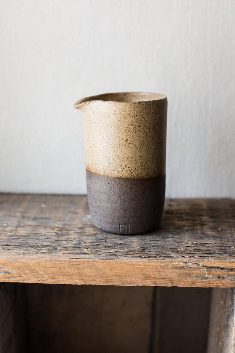 May include: A handmade ceramic creamer with a two-tone design. The top half is a speckled tan color, while the bottom half is a dark brown. The creamer has a small spout for easy pouring and is set on a wooden surface.